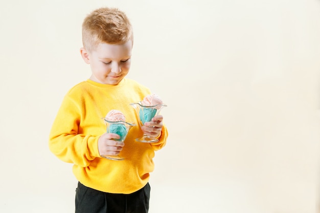 Un hermoso niño pelirrojo de 5 años con un suéter naranja se alza sobre un fondo claro con helado en sus manos