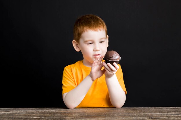 Un hermoso niño pelirrojo con una deliciosa magdalena de chocolate, un niño come una magdalena con placer, dulce comida dañina pero deliciosa en un niño