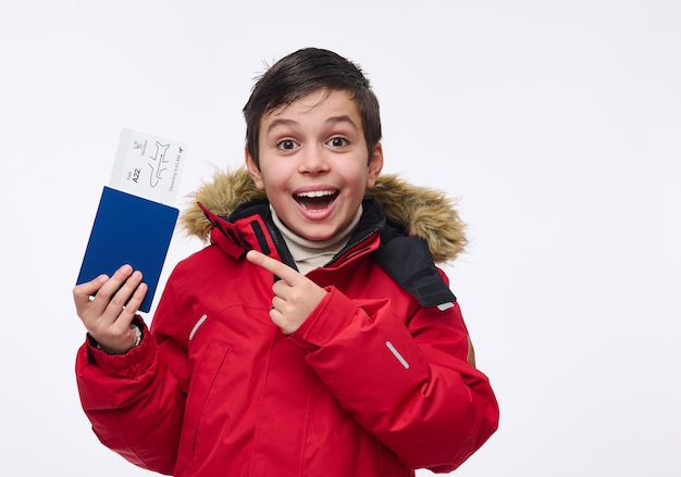 Hermoso niño preadolescente, niño de escuela alegre en parka cálida de color rojo brillante apunta con su dedo a un pasaporte con un boleto y tarjeta de embarque, aislado sobre fondo blanco con espacio publicitario de copia