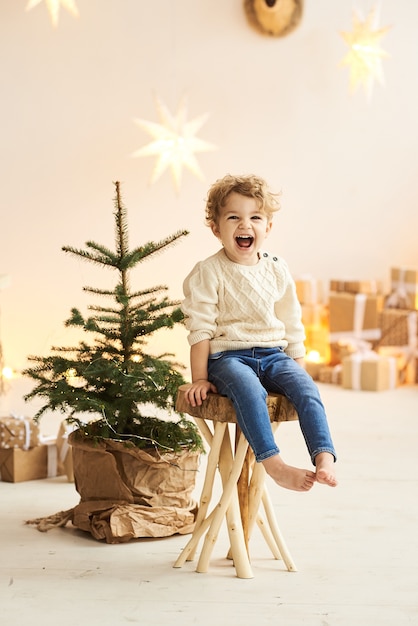 Hermoso niño rizado decorado un árbol de Navidad en una habitación blanca