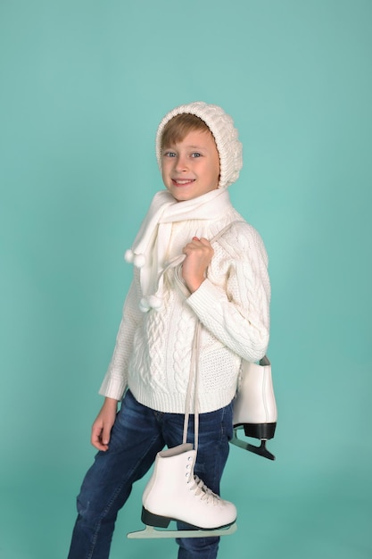 Un hermoso niño rubio feliz con un suéter blanco de punto y un sombrero sosteniendo patines