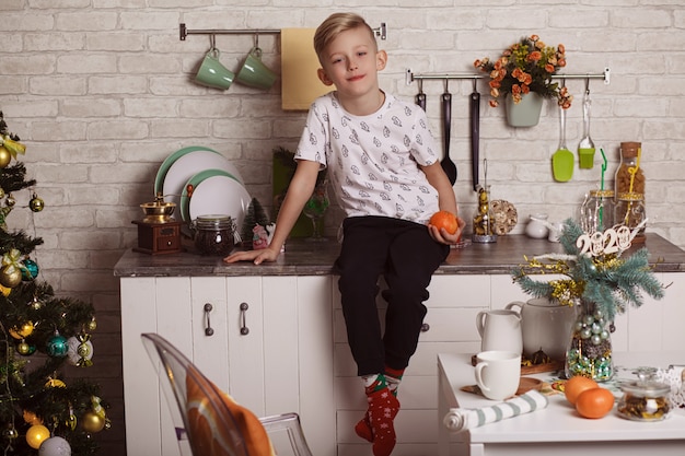 Hermoso niño rubio se sienta en la mesa de la cocina y sostiene una naranja en la mano