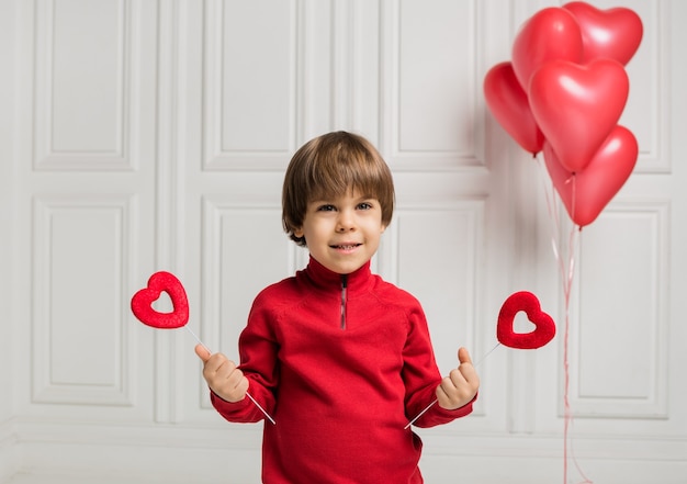 Hermoso niño sosteniendo dos corazones en un palo sobre un fondo blanco con corazones de globos