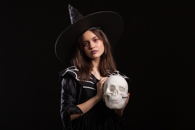 Hermoso niño vestido con un vestido negro para el carnaval de halloween con un cráneo humano en las manos. Chica aislada sobre fondo negro con un disfraz de bruja.