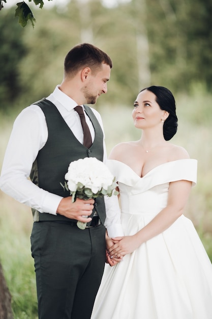 Hermoso novio fuerte con su joven novia bonita en vestido blanco con un ramo de flores blancas a pie del jardín verde