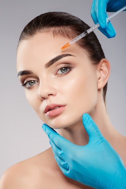 Hermoso paciente con maquillaje desnudo en el fondo del estudio, las manos del médico con guantes azules cerca de la cara del paciente, sosteniendo la jeringa con botex cerca de la cara, mirando a la cámara.