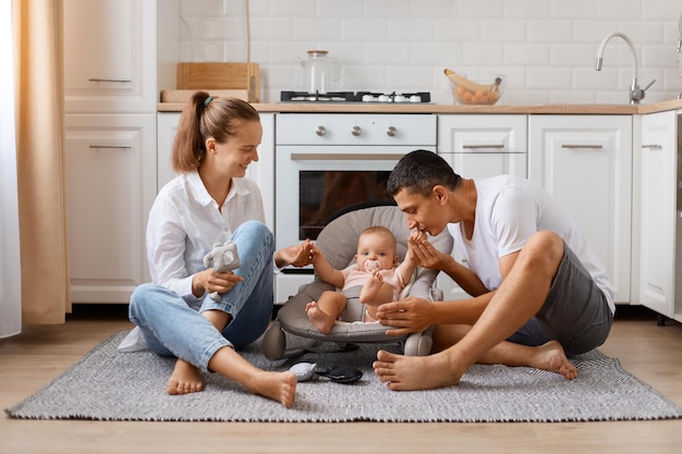 Hermoso padre de familia caucásica, madre e hija sentados en el piso en la acogedora cocina blanca, padres amorosos disfrutando del tiempo con su lindo niño, padre besando la mano del niño.