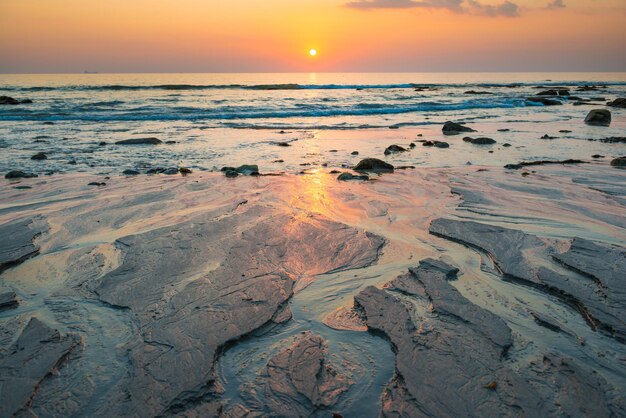 Hermoso paisaje colorido atardecer con playa de arena, sol dorado y piedras en la orilla del mar