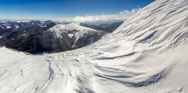 Hermoso paisaje invernal Ladera empinada de la colina de la montaña con nieve blanca y profunda Panorama lejano de la cordillera leñosa que se extiende hasta el horizonte y rayos de sol brillantes en el fondo del espacio de la copia del cielo azul