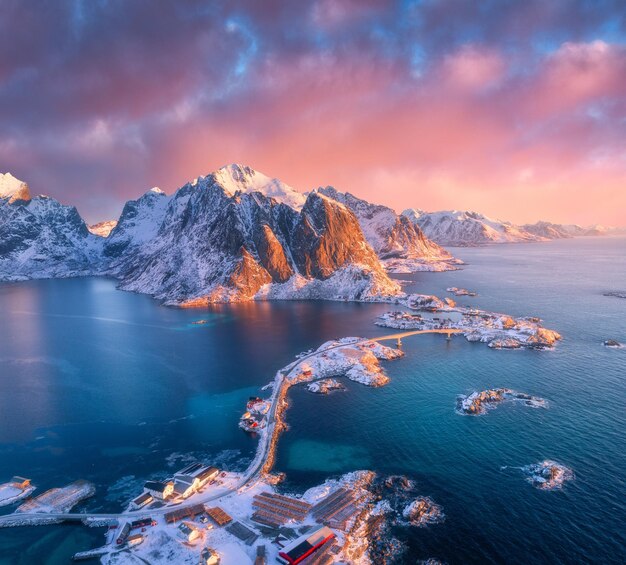 Hermoso paisaje con mar azul montañas nevadas rocas e islas pueblo puente de carretera rorbu y cielo rosa Vista aérea Hamnoy en la nieve al amanecer en invierno en las islas Lofoten Noruega Vista superior