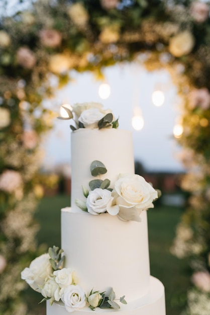 Hermoso pastel de bodas blanco de varios niveles decorado con flores en el fondo de un arco de bodas por la noche