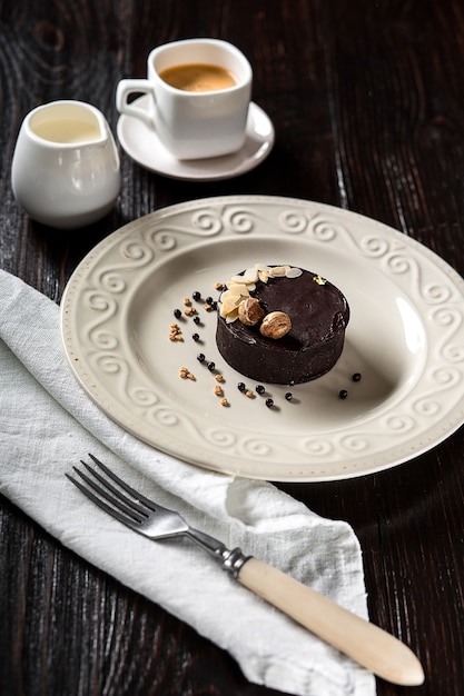 Hermoso pastel de chocolate decorado con nueces doradas sobre un plato blanco y una taza de café sobre un fondo de madera negra