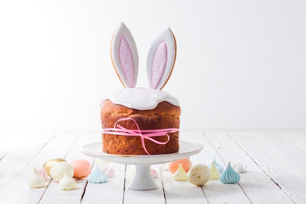 El hermoso pastel de Pascua decorado con orejas de conejo comestibles El pastel envuelto en cinta rosa se encuentra en una bandeja blanca Al lado de la mesa hay huevos de color