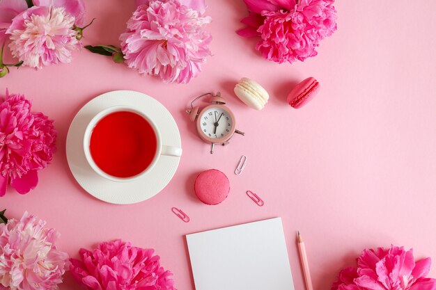 Hermoso plano sentar con bloc de notas, lápiz, reloj despertador, flores de peonía, una taza de té y macarons