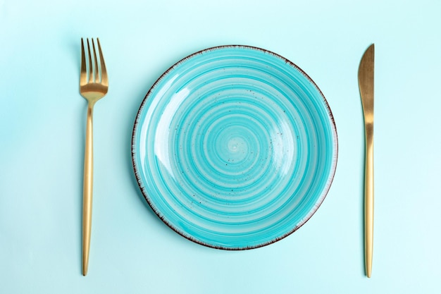 Hermoso plato de cerámica redondo azul vacío con cubiertos dorados, tenedor y cuchillo sobre fondo azul claro Ajuste de la mesa Vista superior Espacio de copia Maqueta Orientación horizontal