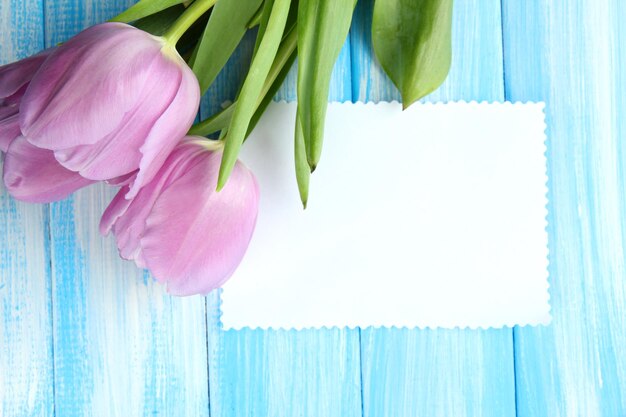 Hermoso ramo de tulipanes morados y tarjeta en blanco sobre fondo de madera azul
