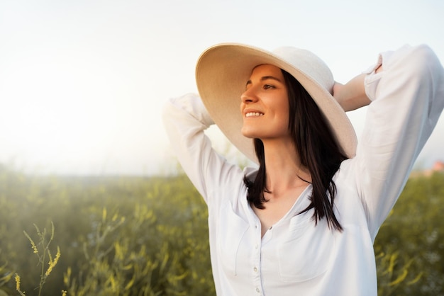 Hermoso retrato de una mujer joven romántica y atractiva con las manos en el sombrero y la camisa blanca soñando mirando hacia el cielo en el fondo del campo de la naturaleza Belleza