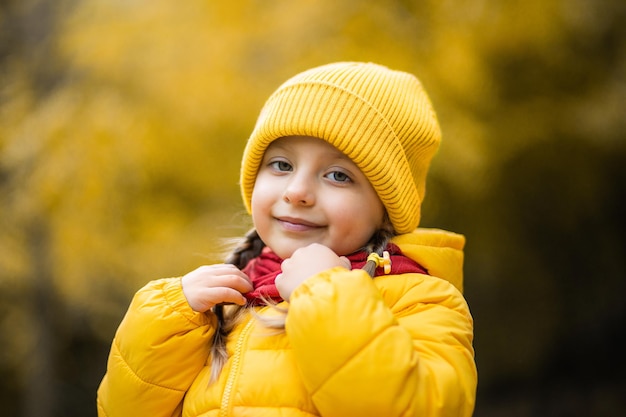 Hermoso retrato de otoño al aire libre de adorable niña de 4 años de edad, vestida con elegante capa y gorra amarilla, tocando su cálida bufanda roja y sonriendo a la cámara.