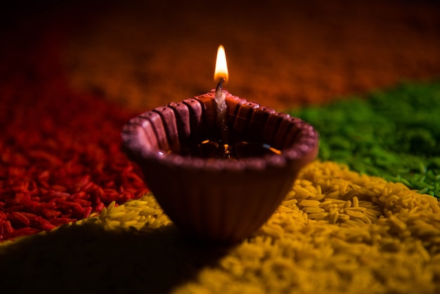 Hermoso saludo de Diwali con Diya o lámpara de aceite de arcilla encendida y dispuesta sobre Rangoli hecha por granos de arroz multicolores, enfoque selectivo