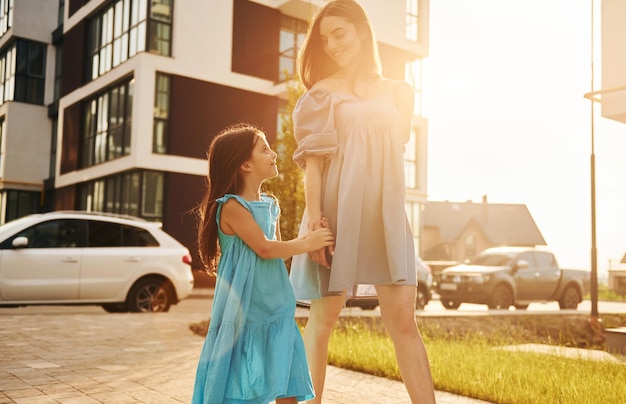 Hermoso sol Madre joven con su pequeña hija caminando cerca de los edificios