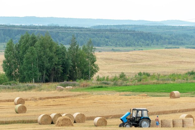 Un hermoso tractor azul se encuentra en un campo con la tapa del radiador abierta Sobrecalentamiento y avería de equipos agrícolas Fardos de heno están esparcidos por el campo