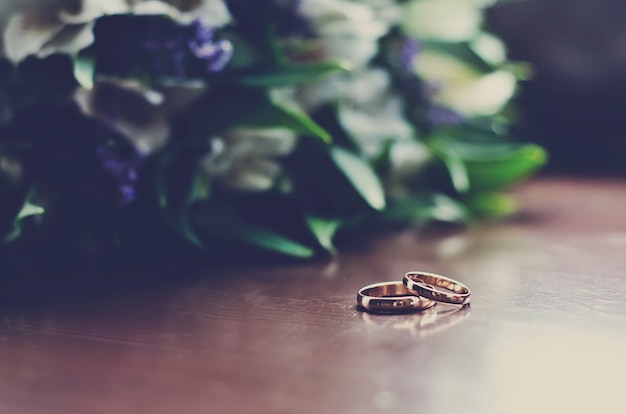 Hermosos anillos de boda se encuentran sobre una superficie de madera con el telón de fondo de un ramo de flores