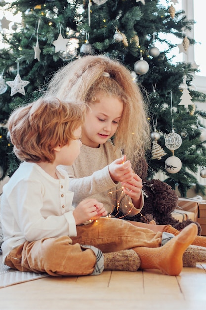 Hermosos hermanos y hermanas juegan con luces y se sientan debajo del árbol de Navidad.