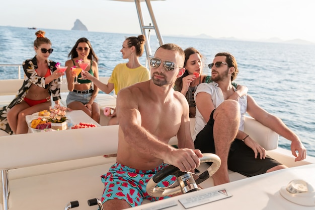 Hermosos jóvenes se relajan en un yate de lujo. Se sientan a la mesa, beben cócteles y comen frutas tropicales. Concéntrese en el apuesto capitán con el torso desnudo que conduce el yate.