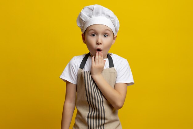 Foto hermosos niños caucásicos de seis o siete años en un uniforme de chef se muestran aturdidos cubriendo su boca con