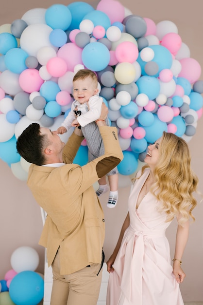 Hermosos padres jóvenes sonríen con su hijo de un año sobre fondo de globos de color rosa y azul.