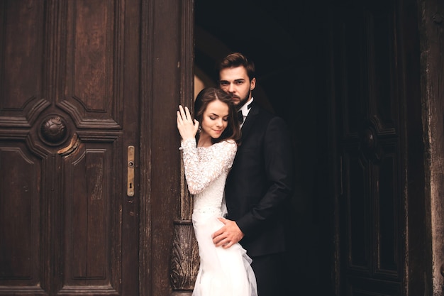 Hermosos recién casados abrazándose cerca de la puerta antigua. Retrato de boda de un elegante novio y una joven novia cerca de la casa antigua en una ciudad europea.