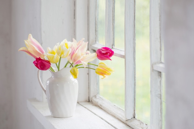 Hermosos tulipanes en florero en el alféizar de la ventana blanca