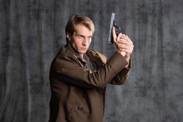 Foto héroe de estilo militar con una pistola en las manos un joven con un largo abrigo marrón y calzones con