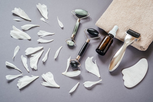 Herramientas antienvejecimiento para el cuidado de la piel: rodillo derma de microagujas, rodillos de masaje jade guasha y frasco de suero con pétalos de peonía blanca sobre fondo gris