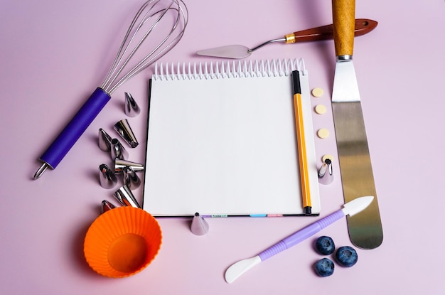 Herramientas para crear y decorar un pastel varias espátulas y boquillas de crema con un cuaderno abierto y un bolígrafo para texto y recetas sobre un fondo morado