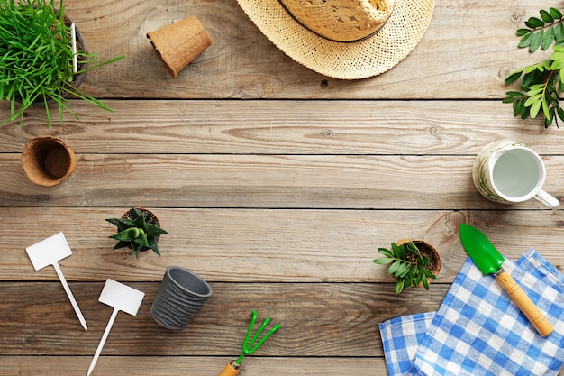 Herramientas de jardinería, flores en maceta, hierba y sombrero de paja sobre fondo de madera vintage.