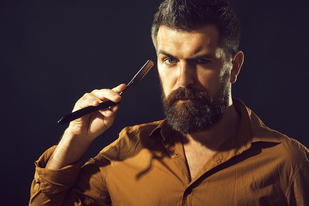 Herramientas para peluquería o afeitado. Peluquero atractivo con barba negra y bigote sostiene una navaja de afeitar cerca de su rostro. Macho sexy guapo barbudo con navaja de afeitar. Hombre sin afeitar dispuesto a afeitarse la barba.