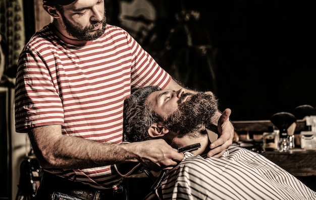 Herrenfriseur rasiert seinen Bart Bärtiger Mann, der in einem Friseursalon in einem Sessel sitzt, während der Friseur seinen Bart mit einem gefährlichen Rasiermesser rasiert