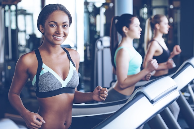 Herrlich und sportlich. Schöne junge fröhliche Afrikanerin in Sportbekleidung, die mit einem Lächeln in die Kamera schaut, während sie mit anderen Frauen auf dem Laufband im Fitnessstudio läuft