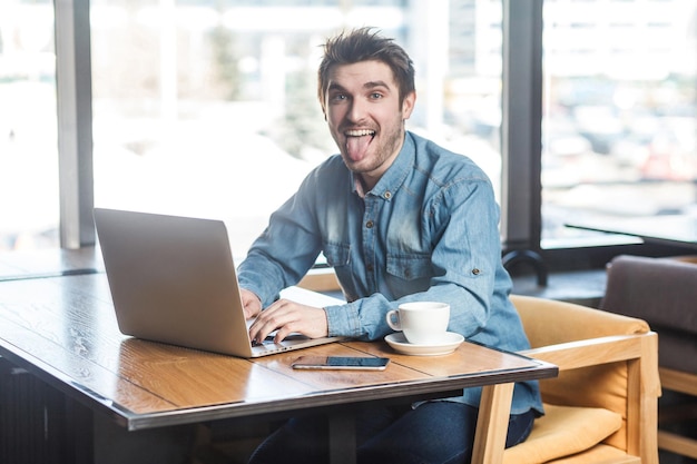 Herumalbern. Porträt eines gutaussehenden, kindischen, positiven, bärtigen jungen Freiberuflers in Blue Jeans-Hemd sitzt im Café und arbeitet am Laptop, zeigt die Zunge heraus und schaut in die Kamera.