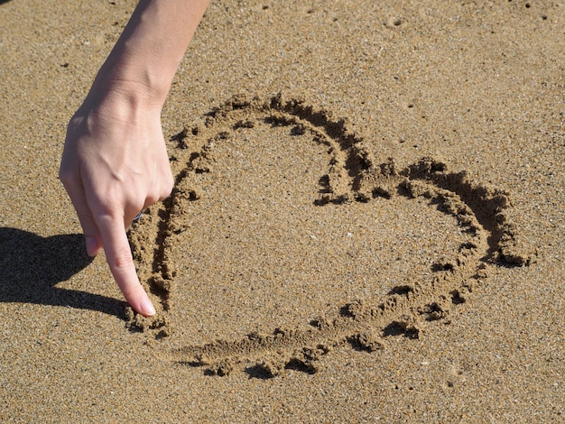 Herz, das einen Finger im Sand zeichnet. Frauenhand.