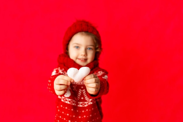 Herz in den Händen eines kleinen Mädchens in der Winterkleidung auf einem roten Hintergrund. Neujahrs- oder Valentinstagskonzept, Platz für Text