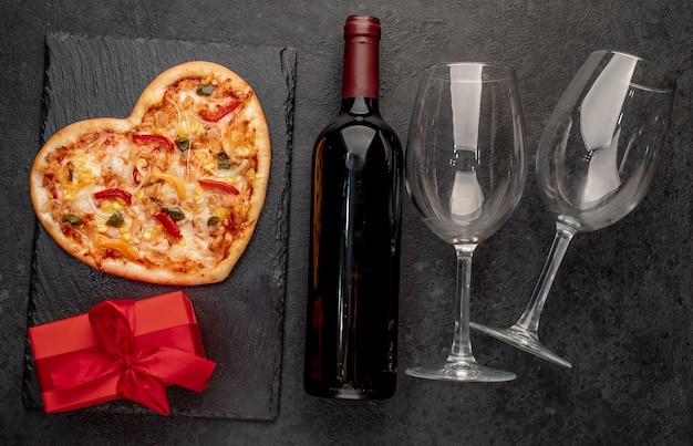 Herzförmige Pizza zum Valentinstag mit einer Flasche Wein und zwei Gläsern auf Schiefer