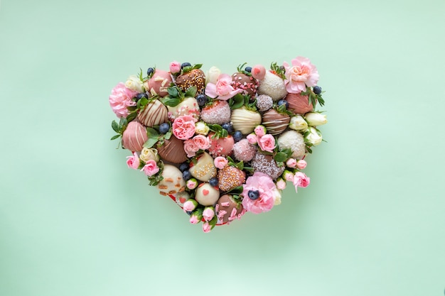 Herzförmige Schachtel mit handgemachten Erdbeeren mit Schokoladenüberzug und verschiedenen Belägen und Blumen als Geschenk am Valentinstag auf grünem Hintergrund