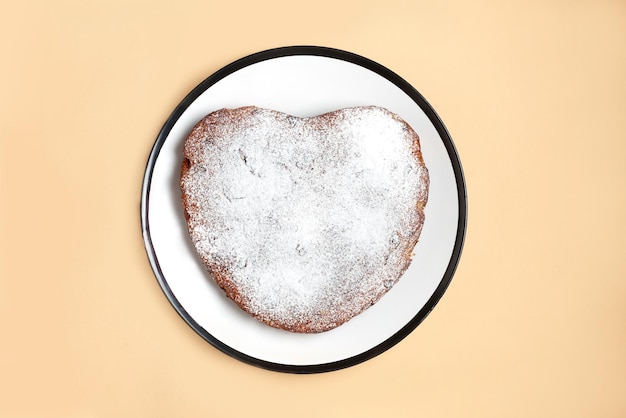 Herzförmiger Kuchen, der nach Hause gepuderten Zuckerkuchen kocht, der auf weißer Platte auf beigem Hintergrund servierfertig ist