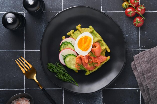 Herzhafte Waffeln Frühstück grüne Waffeln Bärlauch oder Spinat mit Ei Tomaten Lachs auf Holzplatte zum Frühstück auf alten schwarzen Kacheltisch Hintergrund Leichtes Abendessen, Mittag- oder Frühstück Draufsicht