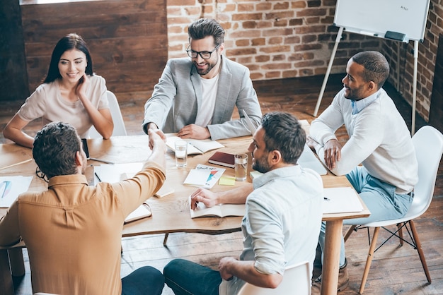 Herzlichen Glückwunsch zu Ihrer Beförderung! Draufsicht auf zwei Männer, die Hände schütteln und lächeln, während ihre Kollegen im Büro am Tisch sitzen table