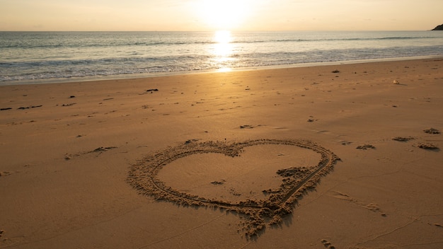 Herzsymbol am Strand, Hand gezeichnetes Herz auf Strandsand über Sonnenuntergang oder Sonnenaufganghimmel schöne helle Naturlandschaft.