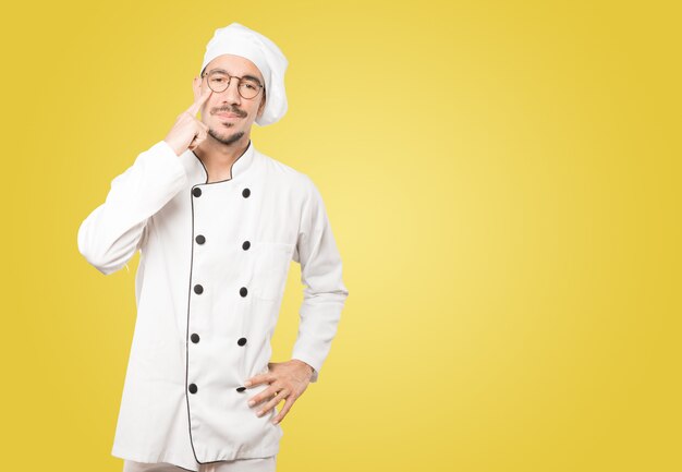Hesitante jovem chef fazendo um gesto de cuidado com a mão apontando para o olho