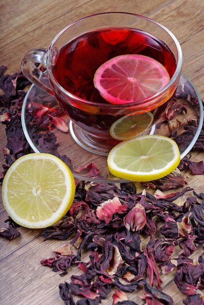 Hibiskustee und Zitrone in einem Glasbecher auf einem Holztisch unter den Rosenblättern und dem trockenen Tee